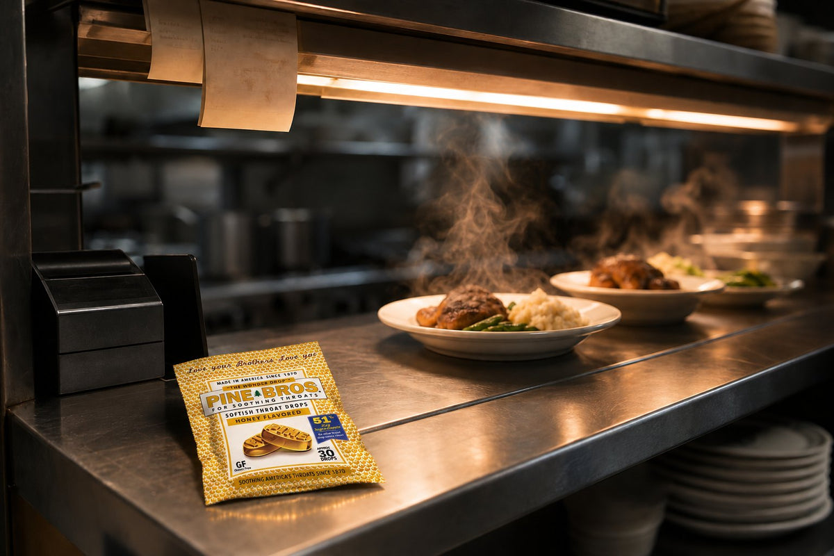 A restaurant kitchen pass window in mid-October, tickets clipped above, steam rising from plates, and a Pine Brothers Honey pouch on the counter edge, warm kitchen light