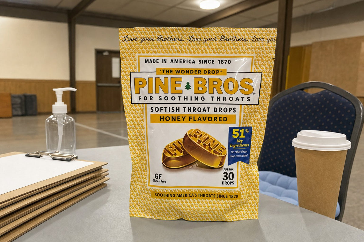 A polling place table set up in a community hall in early November, a stack of clipboards, a hand sanitiser bottle, a coffee cup, a Pine Brothers Real Honey pouch beside a seat cushion, overhead hall light