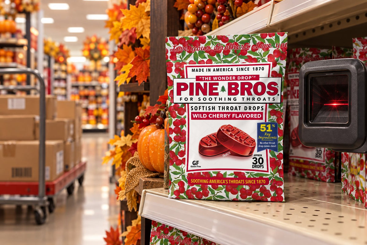 A store aisle on the last week of October, empty cartons on a flatbed cart, shelf tags, a seasonal display endcap, a Pine Brothers Wild Cherry pouch on the shelf edge beside a price scanner, warm retail light