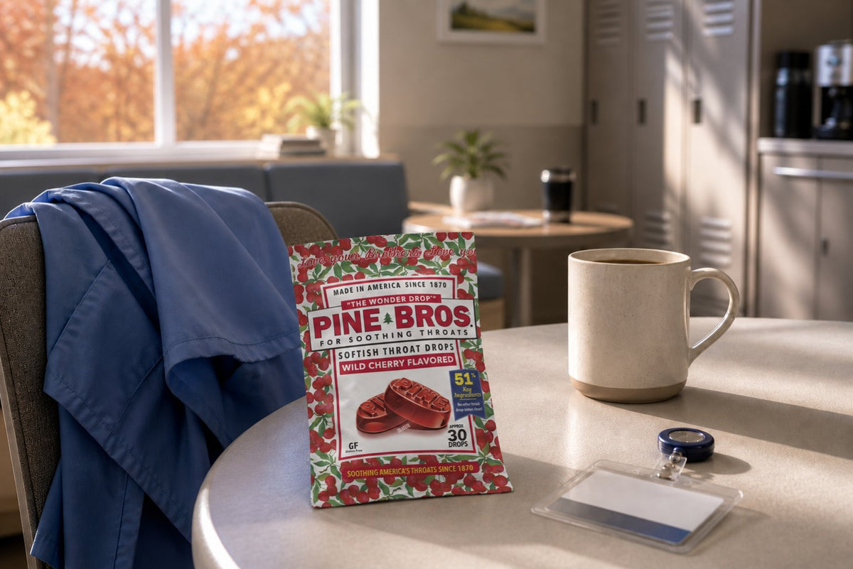 A quiet hospital break room in early fall with a scrub jacket draped over a chair, a name badge, a coffee mug, and a Pine Brothers Wild Cherry pack on the table, soft morning window light