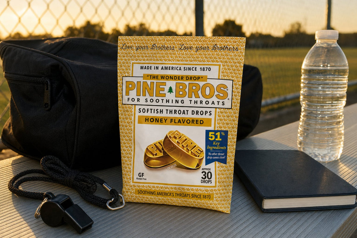 A referee's bag on a bench at an outdoor field on an October evening, a whistle on a lanyard, a rulebook, a bottle of water, a Pine Brothers Real Honey pouch in the side pocket, cold autumn light with long shadows