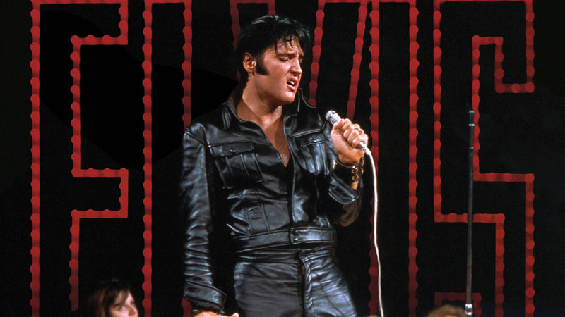 Elvis Presley singing into a microphone in black leather in front of a red-lit ELVIS sign, 1968
